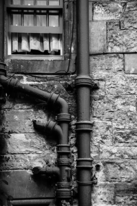 Grayscale image of a sewer drain pipe down the wall 